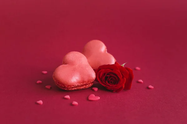 Macarrones en forma de corazón con rosa roja sobre un fondo rojo oscuro, decorados con corazones dulces . — Foto de Stock