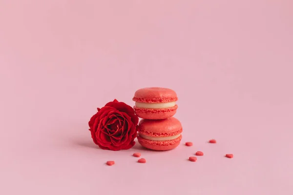 Lekkere franse macarons met rode roos op een roze pastelachtergrond, versierd met lieve hartjes. — Stockfoto