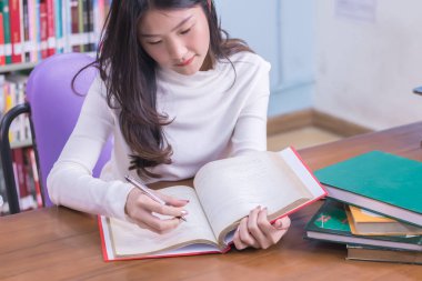Genç ve güzel Asyalı kız öğrenci portresi üniversite kütüphanesinde ders kitabı okuyarak ya da ders kitabı okuyarak arka planda kitap rafları ile sınav yapıyor. Eğitim ve öğrenme kavramı