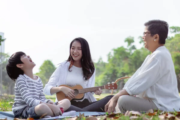 Happy Asian family has leisure in the park on the weekend or holiday. Healthy grandmother and lovely son enjoy daughter or mother play guitar. Family generation relationship or bonding photo concept