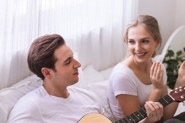 Lovely young caucasian couple or lover on bed together. Man play guitar to woman in dreamy romantic moment in bedroom. Photo of married relationship or bonding between boyfriend and girlfriend