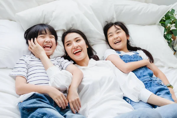 Happy Asian family smiling mother, son & daughter on bed in bedroom. Mom, son & daughter have fun together at home. Love relationship or bonding between mum and children. People lifestyle concept
