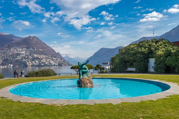 Switzerland - March 10, 2019: A fountain by Lake Lugano with a beautiful statue in the middle of blue water in Ticino canton of Switzerland — Stock Photo, Image