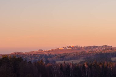 Köyle birlikte kırsal alanda gün batımı - ön planda orman, tepedeki ufukta kilise ve çeşitli binalar, turuncu renkli gökyüzü