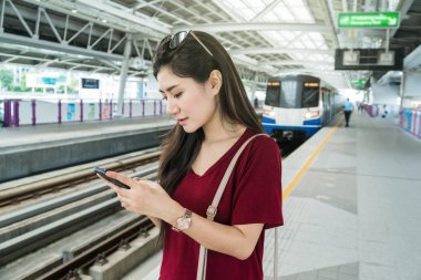 Asyalı kadın yolcu seyahat büyük şehirde Bts Skytrain raylar veya Mrt metro akıllı cep telefonu üzerinden sosyal ağ kullanarak rahat takım elbise ile