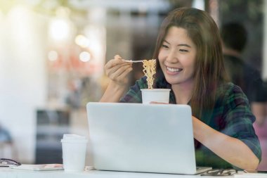 Portrait of Asian businesswoman in casual suit eating noodles with happiness action in rush hour at desk beside glass in modern office. Business work hard concept clipart