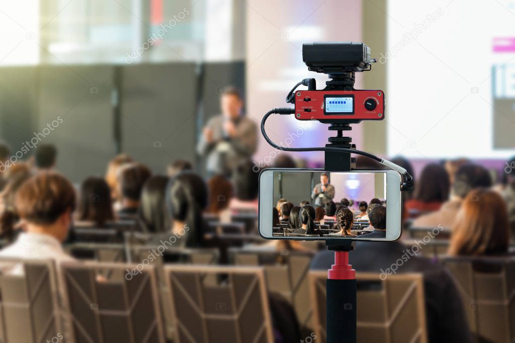 Closeup smart mobile phone taking Live over Speakers on the stage with Rear view of Audience in the conference hall or seminar meeting, technology live streaming and broadcast concept