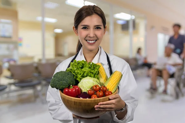 病院の背景 健康食品のコンセプトの抽象的な写真を木製のボウルにミックス新鮮な野菜を保持している美しいアジアの若い医師の肖像画がぼやけています — ストック写真