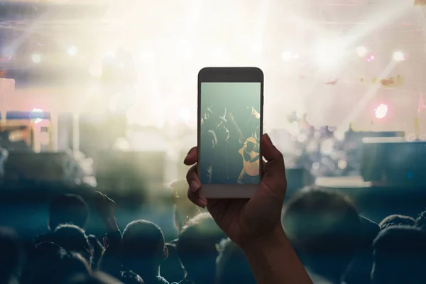 Female hand holding mobile smart phone taking video record or Live stream of Concert crowd with super star songer and silhouettes of Music fanclub with show hand action, musical and concert concept