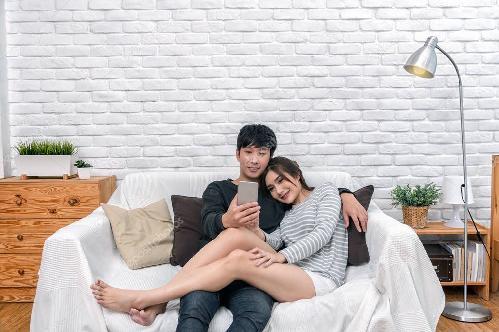 Happy Asian couple sitting and using the digital smart mobile phone to selfie over the sofa of living room at modern home, Couple successfull and lifestyle concept