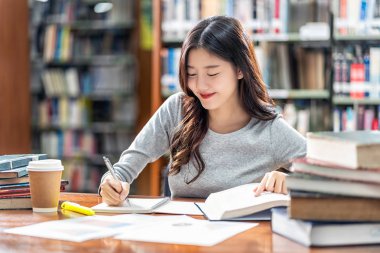 Sıradan takım elbiseli Asyalı genç Öğrenci okuma ve kitap raf arka plan üzerinde ahşap masa üzerinde çeşitli kitap ve sabit ile üniversite veya kolaj kütüphanede ev ödevi yapıyor, Okula geri