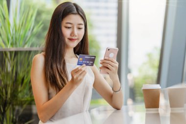 Genç Asyalı bir kadın kahve dükkanında online alışveriş için cep telefonu kullanıyor ya da pencere aynası, teknoloji para cüzdanı ve online ödeme konsepti, kredi kartı modeli