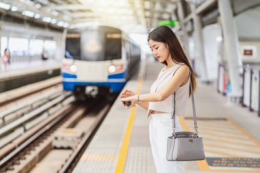 Genç Asyalı kadın yolcu akıllı cep telefonuyla müzik dinliyor ve metro istasyonunda El saatiyle trene bakıyor. Japonca, Çince, Kore yaşam tarzı, banliyö ve ulaşım konsepti.