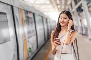 Genç Asyalı kadın yolcu metro istasyonunda akıllı cep telefonuyla müzik kullanıyor ve dinliyor, Japonca, Çince, Kore yaşam tarzı, boş ve günlük yaşam, banliyö ve ulaşım kavramı