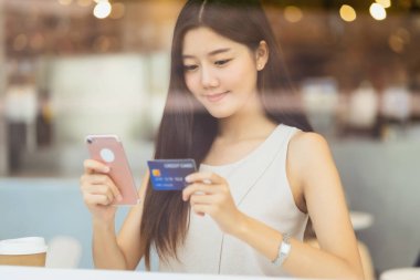 Genç Asyalı bir kadın kahve dükkanında online alışveriş için cep telefonu kullanıyor ya da pencere aynası, teknoloji para cüzdanı ve online ödeme konsepti, kredi kartı modeli