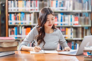 Sıradan takım elbiseli Asyalı genç Öğrenci ev ödevi yapıyor ve üniversite veya kolaj kütüphanesinde çeşitli kitap ve kitap raf arka plan üzerinde sabit ile teknoloji dizüstü bilgisayar kullanarak, Okula Geri