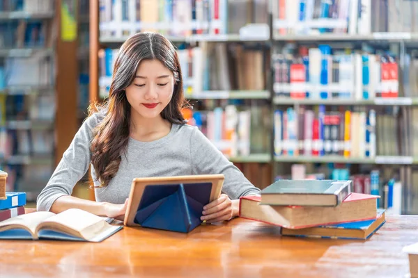 身着休闲装的亚洲青年学生 穿着休闲装 在大学或学院的图书馆里做作业 使用技术词汇 书架上有各种各样的书籍和固定的书架 回到学校 — 图库照片