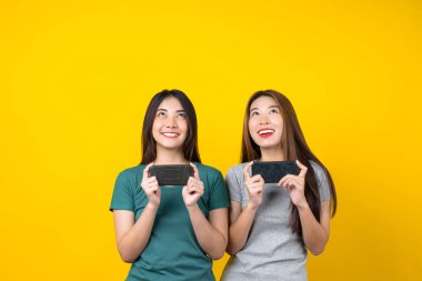 İki Mutluluk Asyalı gülümseyen genç bayan oyuncu akıllı cep telefonu kullanıyor ve izole edilmiş sarı arka plan, yaşam tarzı ve hobi konseptiyle oyun oynuyor.