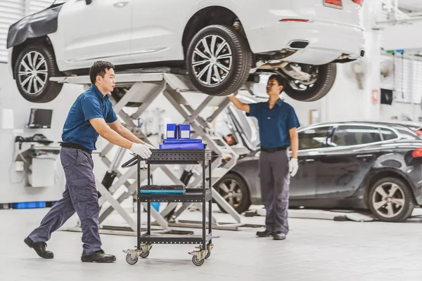Asian mechanic Push cart with car equipment over colleague Checking and repairing the car in maintenance service center which is a part of showroom, technician or engineer professional work