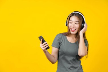Mutlu Asyalı gülümseyen, teknoloji kablosuz kulaklık takan genç bir kadın akıllı cep telefonuyla müzik dinlemek için izole edilmiş sarı arka plan, yaşam tarzı ve hobi konsepti ile boş zaman.