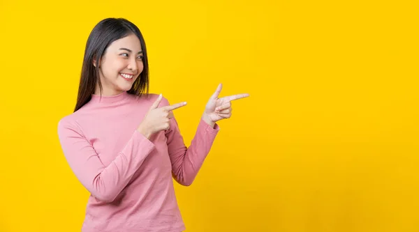 Asian smiling young woman finger Pointing to advertise, banner or showing on isolated yellow color background, wearing winter cozy sweater indoors studio, happiness and fun, copy space concept