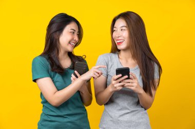İki Mutluluk Asyalı gülümseyen genç bayan oyuncu akıllı cep telefonu kullanıyor ve izole edilmiş sarı arka plan, yaşam tarzı ve hobi konseptiyle oyun oynuyor.