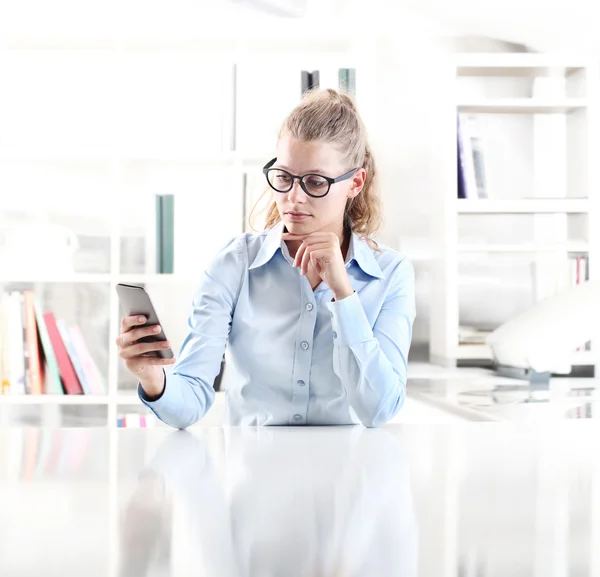 Mujer sentada en el escritorio con el teléfono celular en la mano — Foto de Stock