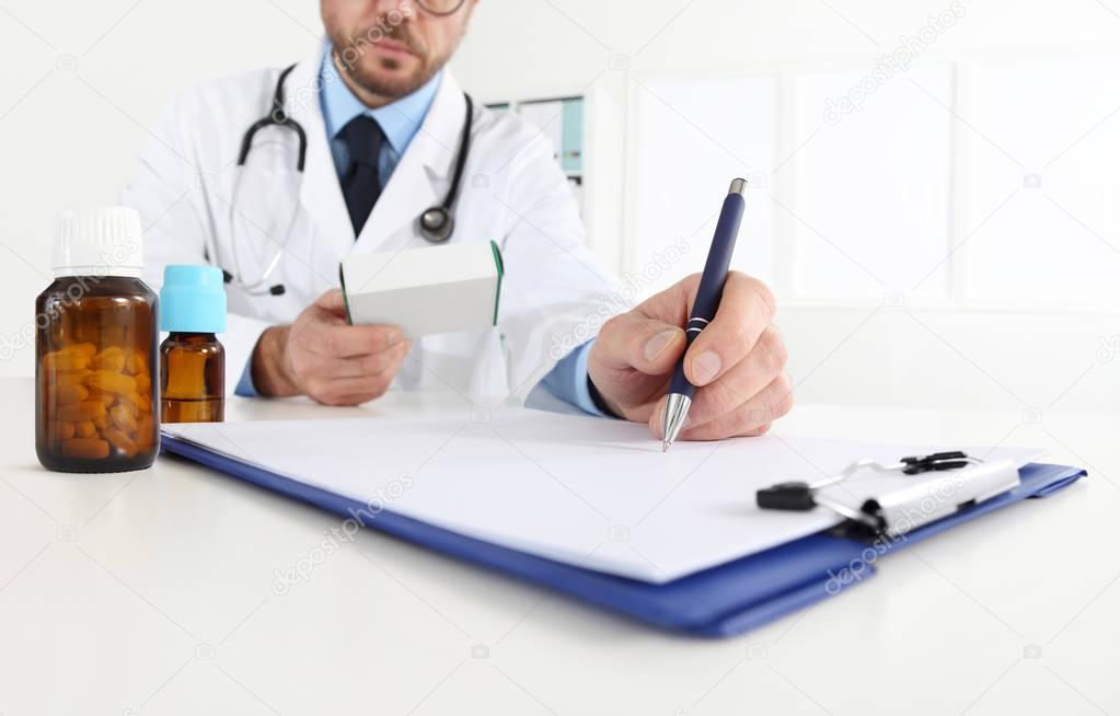 doctor writing RX prescription in medical office with drugs on d