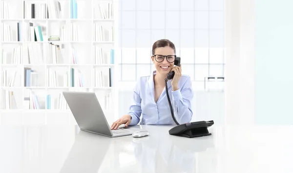 Mujer sonriente en la oficina hablar por teléfono y utilizar la computadora s — Foto de Stock