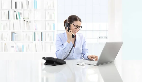 Mujer sonriente en la oficina hablar por teléfono y utilizar la computadora s — Foto de Stock