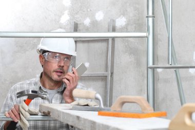 Erkek işi, cep telefonu olan profesyonel inşaat işçisi iskeledeki sıva aletleri, güvenlik başlığı, eldivenler ve koruyucu gözlük. iç inşaat alanının arka planında