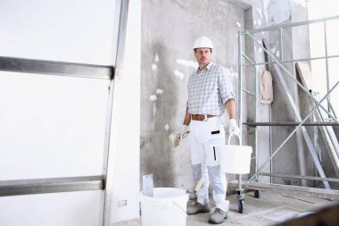 Sıvacı adam bir kova tutuyor ve iç inşaat alanına başvurmadan önce duvarı ıslatıyor miğfer ve koruyucu eldivenler giyiyor, arka planda merdiven ve iskele var.