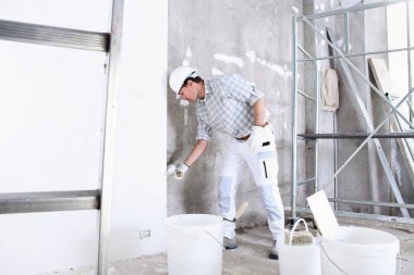 İnşaat alanının duvarını alçılayan alçı işindeki adam miğfer ve koruyucu eldivenler giyer, arka planda merdiven ve iskele olur.