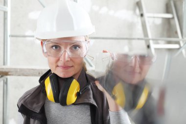 Düz ruh seviyesinde çalışan kadın inşaatçı, kask takıyor, güvenlik gözlüğü takıyor ve kulaklık takıyor, iç yapı iskelesi arka planında çalışıyor.