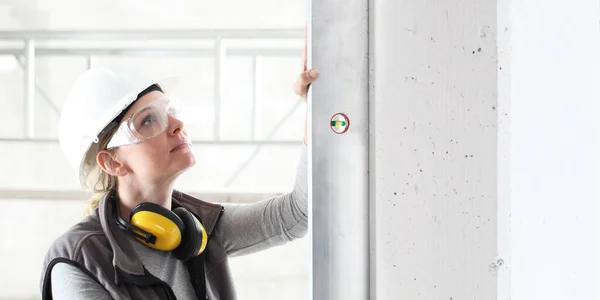 woman worker looks at the spirit level checks the wall with hard hat, glasses and ear protection headphones, white wall with copy space