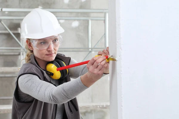 woman construction worker work with meter tape and pencil, measure wall in interior building site, wearing helmet, glasses and hearing protection headphones