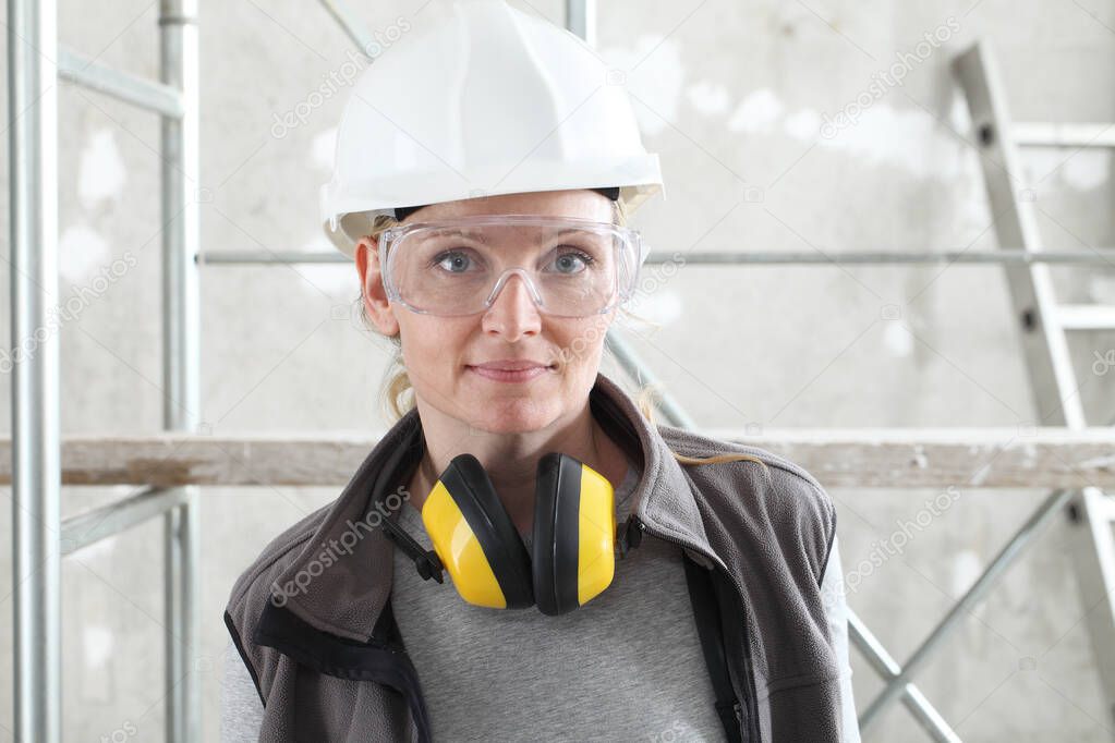 woman worker portrait wearing helmet, safety glasses and hearing protection headphones, scaffolding interior construction site background