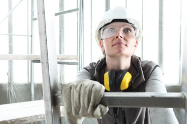 smiling woman construction worker builder on ladder wearing white helmet and hearing protection headphones on interior site building background clipart