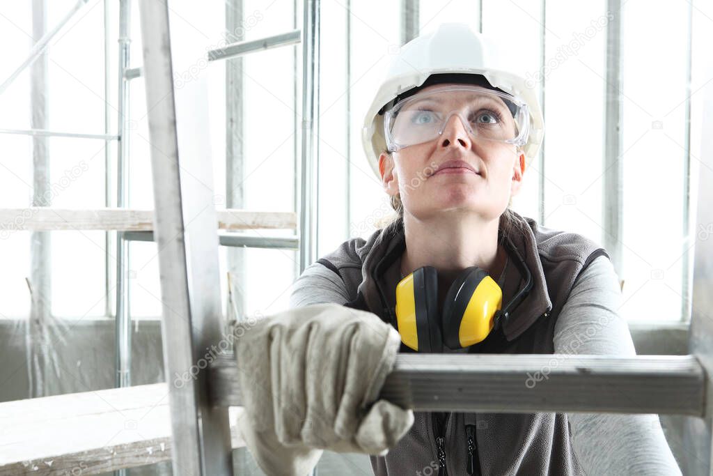 smiling woman construction worker builder on ladder wearing white helmet and hearing protection headphones on interior site building background