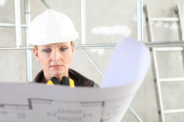 建筑工人中的女性建筑工人在室内背景下看着蓝图 戴着头盔 戴着听觉保护耳机 — 图库照片