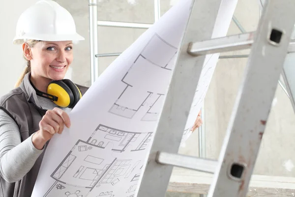 smiling woman construction worker builder looking at bluprint, wearing helmet and hearing protection headphones in building site indoors background
