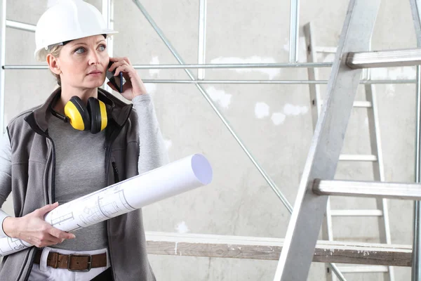 建筑工人中的女建筑工人 她们会讲手机 手握蓝图 头戴头盔 戴着耳机 在建筑工地的室内背景音乐 — 图库照片