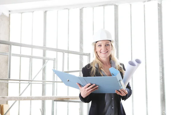 smiling woman architect or construction engineer wear helmet and holds folder and blueprint inside a building site with windows and scaffolding in the background