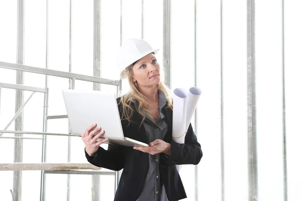 带着电脑笔记本电脑的女建筑师或建筑工程师戴上头盔 并在有窗户和脚手架的建筑工地内放置图纸 — 图库照片