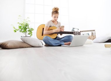 Oturma odasında cep telefonuyla oturan ve bilgisayarda gitar çalan genç bir kadın.