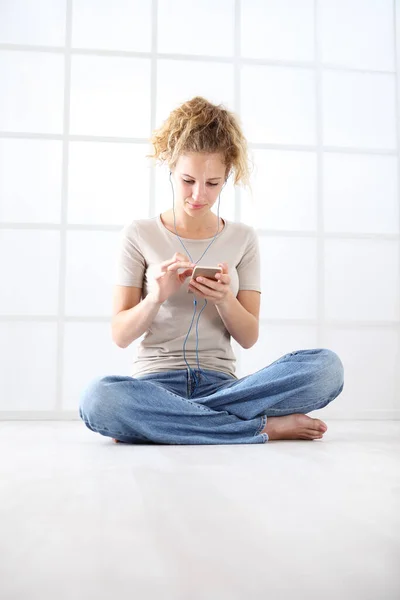 拿着手机的年轻女人坐在地板上 穿着休闲装 一头卷曲的长长的红头发 白色的背景隔离在一起 — 图库照片