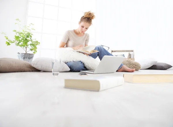 young woman sitting in living room reading a book with your computer nearby, stay at home concept