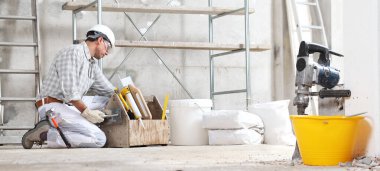 Sıvacı inşaat işçisi alet çantasıyla çalışıyor. Eldiven takıyor, şapka takıyor ve inşaat alanında inşaat iskelesi olan koruyucu gözlükler takıyor. Kova, çuval ve kaya matkabı