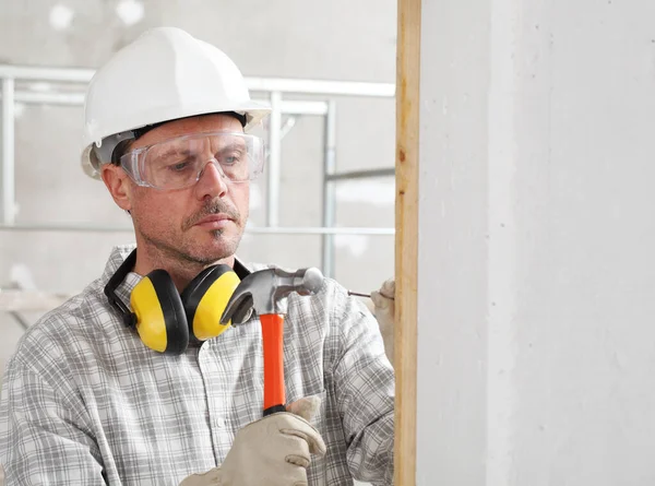 建筑工人用铁锤钉把木板钉在墙上 戴着手套 硬帽子 眼镜和听觉保护耳机 在室内建筑工地 — 图库照片