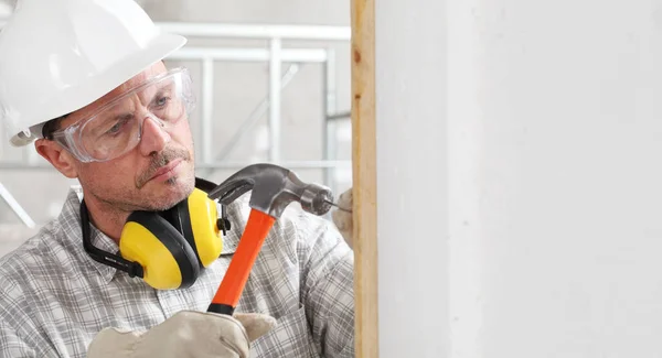 建筑工人用铁锤钉把木板钉在墙上 戴着手套 硬帽子 眼镜和听觉保护耳机 在室内建筑工地 — 图库照片